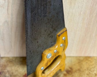 Könnte beinhalten: Vintage Handsäge mit Holzgriff und Metallklinge. Die Klinge ist mit dem "Disston"-Logo und der Modellnummer "D-23" gekennzeichnet. Der Griff ist gelb mit silbernen Nieten. Die Säge ist für die Holzbearbeitung konzipiert.