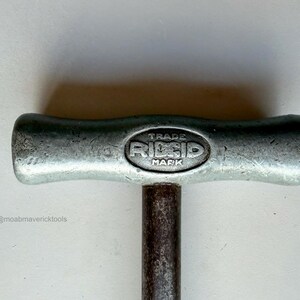 May include: Close-up of a vintage metal tool handle with the text "TRADE RIDGID MARK" embossed on the top. The handle is silver-colored with a dark, cylindrical shaft. The tool appears to be well-used.