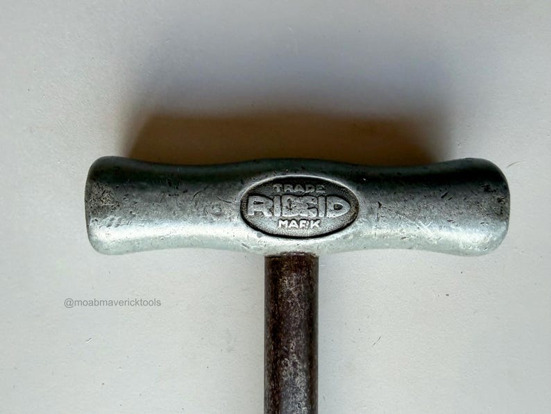 May include: Close-up of a vintage metal tool handle with the text "TRADE RIDGID MARK" embossed on the top. The handle is silver-colored with a dark, cylindrical shaft. The tool appears to be well-used.