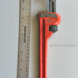 May include: A red and black Ridgid 10-inch pipe wrench with a ruler for scale. The wrench is labeled "HEAVY DUTY" and has a jaw opening. The ruler is marked in inches.