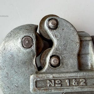 May include: Close-up of a vintage silver-colored pipe cutter tool. The tool has a curved design with a cutting wheel and a spring mechanism. The text "NO 1 & 2" is visible on the tool.