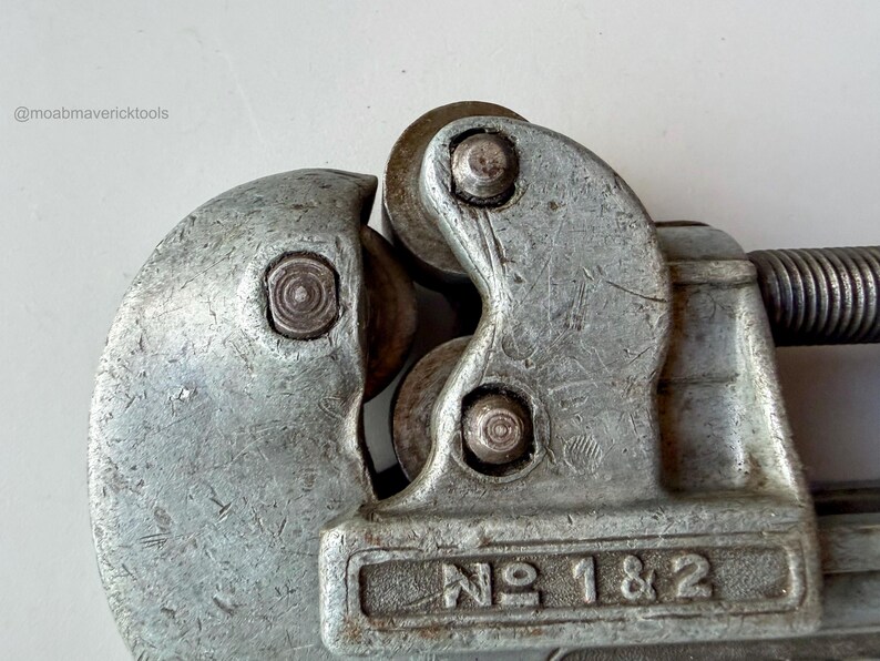 May include: Close-up of a vintage silver-colored pipe cutter tool. The tool has a curved design with a cutting wheel and a spring mechanism. The text "NO 1 & 2" is visible on the tool.