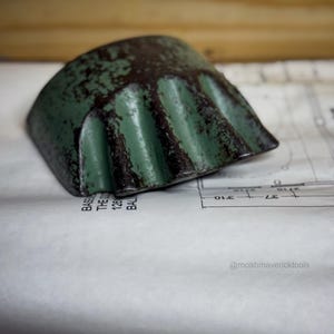 May include: A weathered, green metal tool with a curved, ridged edge. The tool has a distressed appearance with dark brown accents. It rests on a white surface with faint printed lines and text. The tool's design suggests it may be used for shaping or forming materials.