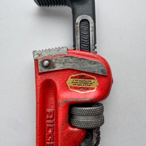 May include: A red and black Ridgid pipe wrench. The wrench has a red handle with the brand name "Ridgid" printed on it. The jaws and head are black. A sticker on the wrench reads "If this Housing ever Breaks or Distorts we will replace it Free."