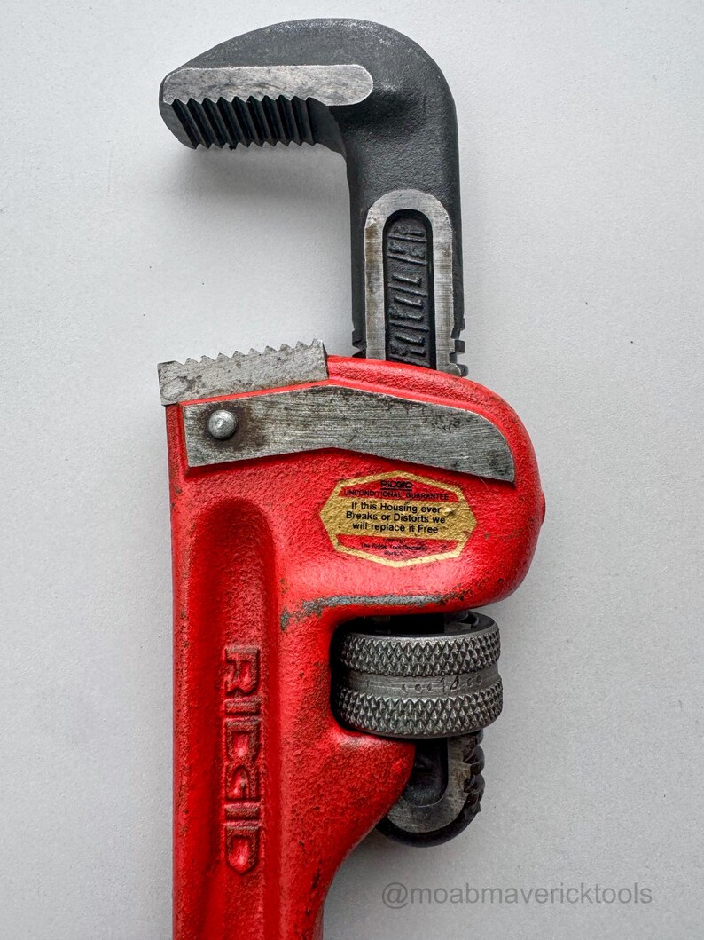 May include: A red and black Ridgid pipe wrench. The wrench has a red handle with the brand name "Ridgid" printed on it. The jaws and head are black. A sticker on the wrench reads "If this Housing ever Breaks or Distorts we will replace it Free."