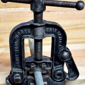 May include: A vintage black Erie Tool Works pipe vise. The vise is made of cast iron and has a screw mechanism for clamping pipes. The text "ERIE TOOL WORKS" and "ERIE PLUS U.S.A." are visible on the frame. A metal pipe is clamped in the vise.