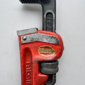 May include: A red and black RIDGID pipe wrench. The wrench has a textured red handle and a black head with serrated jaws. The text "RIDGID" is visible on the handle. The wrench is a tool used for gripping and turning pipes.
