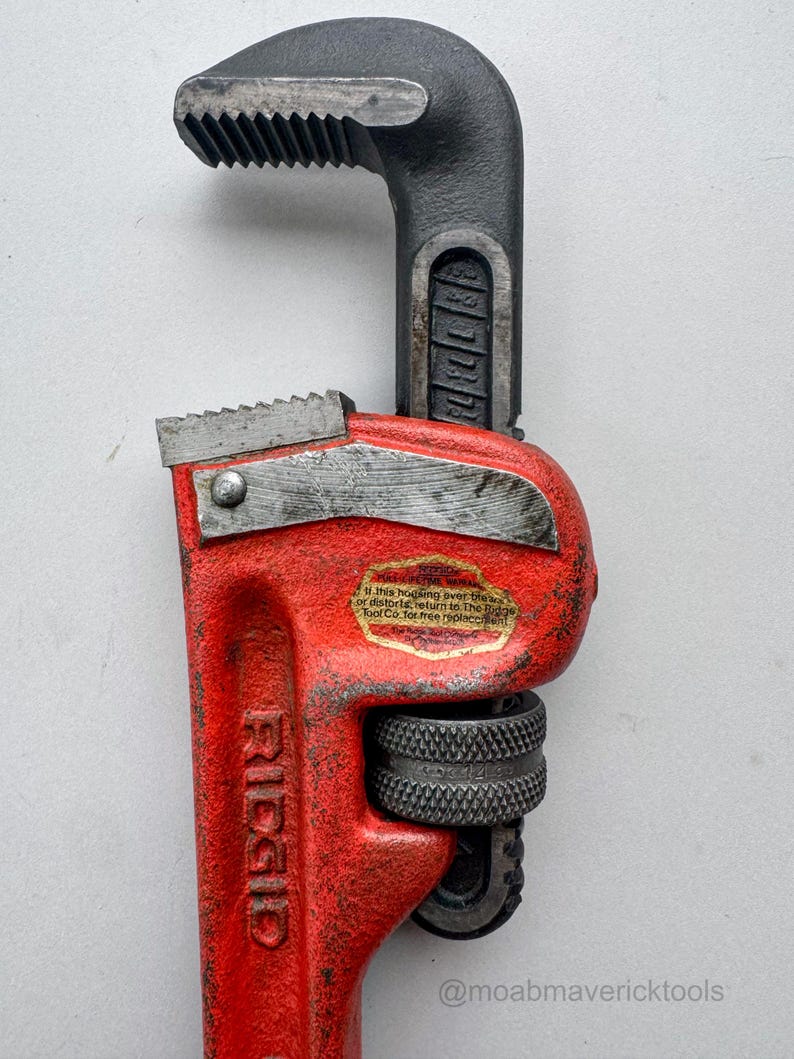 May include: A red and black RIDGID pipe wrench. The wrench has a textured red handle and a black head with serrated jaws. The text "RIDGID" is visible on the handle. The wrench is a tool used for gripping and turning pipes.