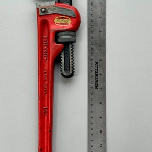 May include: A red Ridgid 14-inch heavy-duty pipe wrench with a black jaw, next to a ruler marked in inches. The wrench has a textured handle and a label. The ruler is marked with the Pittsburgh brand.