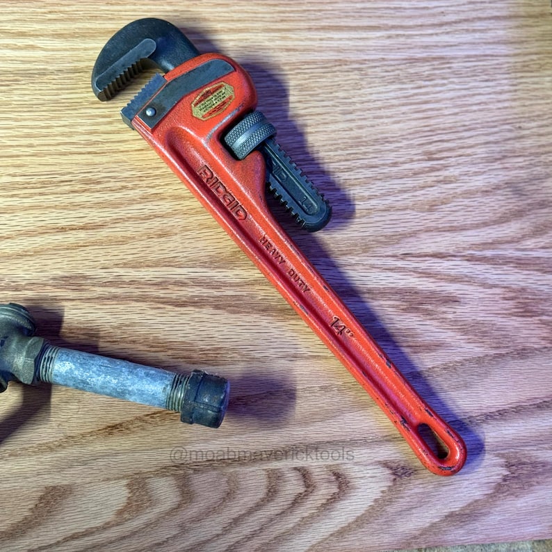 May include: An orange Ridgid heavy-duty pipe wrench, 14 inches long, with a black adjustable jaw and handle. A section of pipe is visible in the lower left corner. The wrench is on a wooden surface.