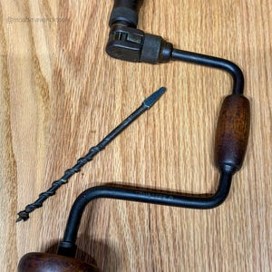 May include: A vintage hand drill with a wooden handle and a black metal frame. A separate drill bit is also present. The drill is lying on a wooden surface. The text "CRUSADER" is visible on the drill.