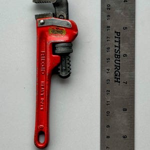 May include: A red Ridgid Heavy Duty pipe wrench with a black adjustable jaw. A metal ruler is next to the wrench. The wrench is approximately 8 inches long.