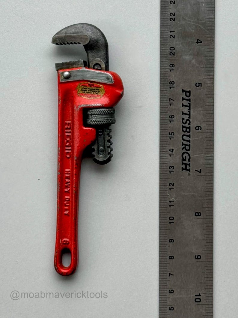May include: A red Ridgid Heavy Duty pipe wrench with a black adjustable jaw. A metal ruler is next to the wrench. The wrench is approximately 8 inches long.