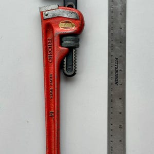 May include: A red Ridgid heavy-duty pipe wrench, 14 inches long, with a black head and adjustable jaw. A metal ruler is placed next to the wrench for scale. The wrench has the text "RIDGID HEAVY DUTY 14" on the handle.