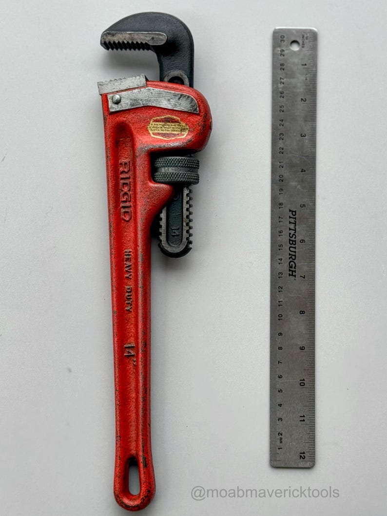 May include: A red Ridgid heavy-duty pipe wrench, 14 inches long, with a black head and adjustable jaw. A metal ruler is placed next to the wrench for scale. The wrench has the text "RIDGID HEAVY DUTY 14" on the handle.