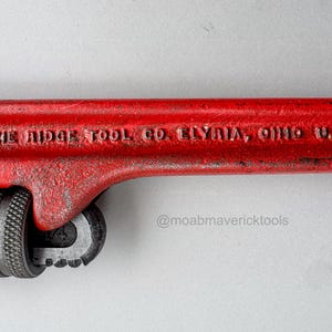May include: A close-up of a red pipe wrench with the text "THE RIDGE TOOL CO. ELYRIA, OHIO U.S.A." The wrench has a textured handle and adjustable jaws. The tool is designed for gripping and turning pipes.