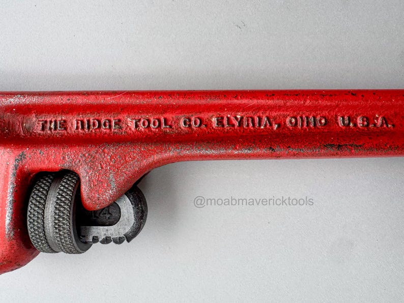 May include: A close-up of a red pipe wrench with the text "THE RIDGE TOOL CO. ELYRIA, OHIO U.S.A." The wrench has a textured handle and adjustable jaws. The tool is designed for gripping and turning pipes.