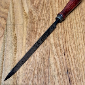 May include: A woodworking rasp file with a dark wooden handle and a black metal blade. The rasp file is lying on a wooden surface. The handle is a dark reddish-brown color.