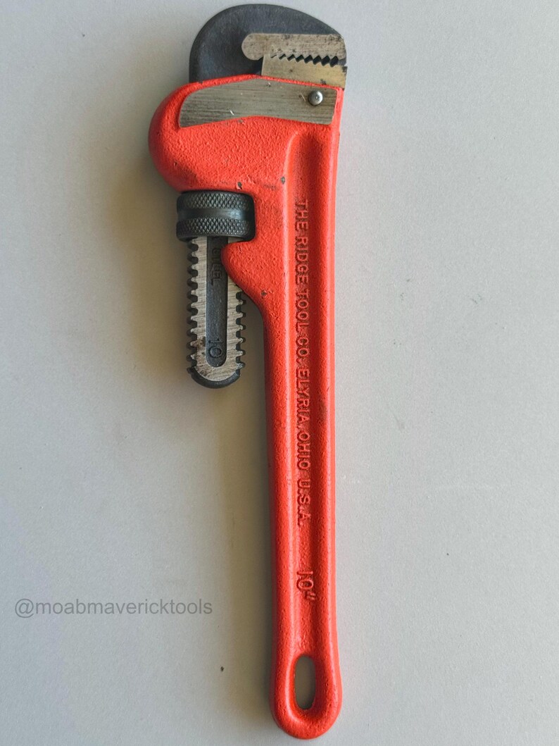 May include: A bright red 10-inch pipe wrench with a black jaw and handle. The wrench has the text "THE RIDGE TOOL CO. ELYRIA, OHIO U.S.A." printed on the handle. The wrench is designed for gripping and turning pipes.