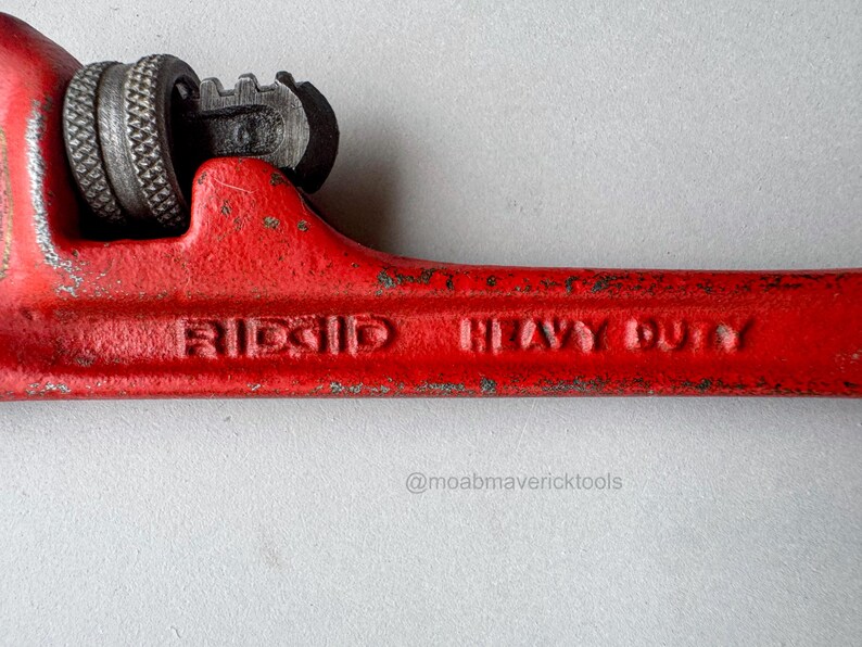 May include: Close-up of a red RIDGID HEAVY DUTY pipe wrench. The wrench has a textured grip and adjustable jaws. The text "RIDGID HEAVY DUTY" is embossed on the handle. The wrench is a tool for plumbing and construction.