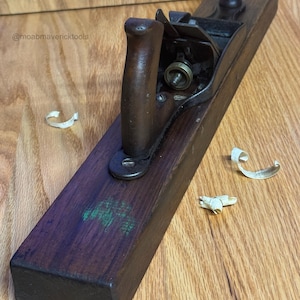 May include: A vintage wooden hand plane with a dark brown finish. The plane has a metal blade and a wooden handle. Wood shavings are scattered around the plane. The text "@moabmavericktools" is visible.