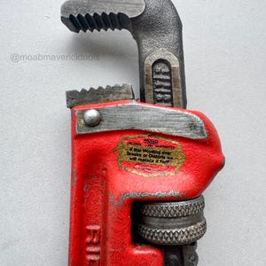 May include: Close-up of a red and gray pipe wrench. The wrench has a red handle and a gray head with serrated jaws. The text "RIDGID" is visible on the handle. The wrench also has a sticker with text that reads "UNCONDITIONAL GUARANTEE".