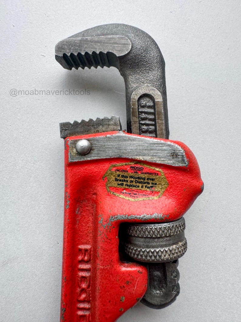 May include: Close-up of a red and gray pipe wrench. The wrench has a red handle and a gray head with serrated jaws. The text "RIDGID" is visible on the handle. The wrench also has a sticker with text that reads "UNCONDITIONAL GUARANTEE".