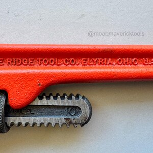 May include: Close-up of a red pipe wrench with the text "THE RIDGE TOOL CO. ELYRIA, OHIO U.S.A." The wrench has a black adjustable jaw and a textured handle. The number "10" is visible on the jaw.