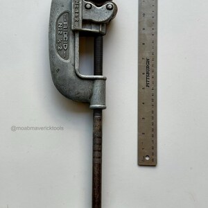 May include: A vintage RIDGID No. 2 pipe cutter, with a silver-colored body and a dark, textured handle. A Pittsburgh ruler, marked in inches, is positioned next to the cutter. The cutter is designed for pipes up to 2 inches.