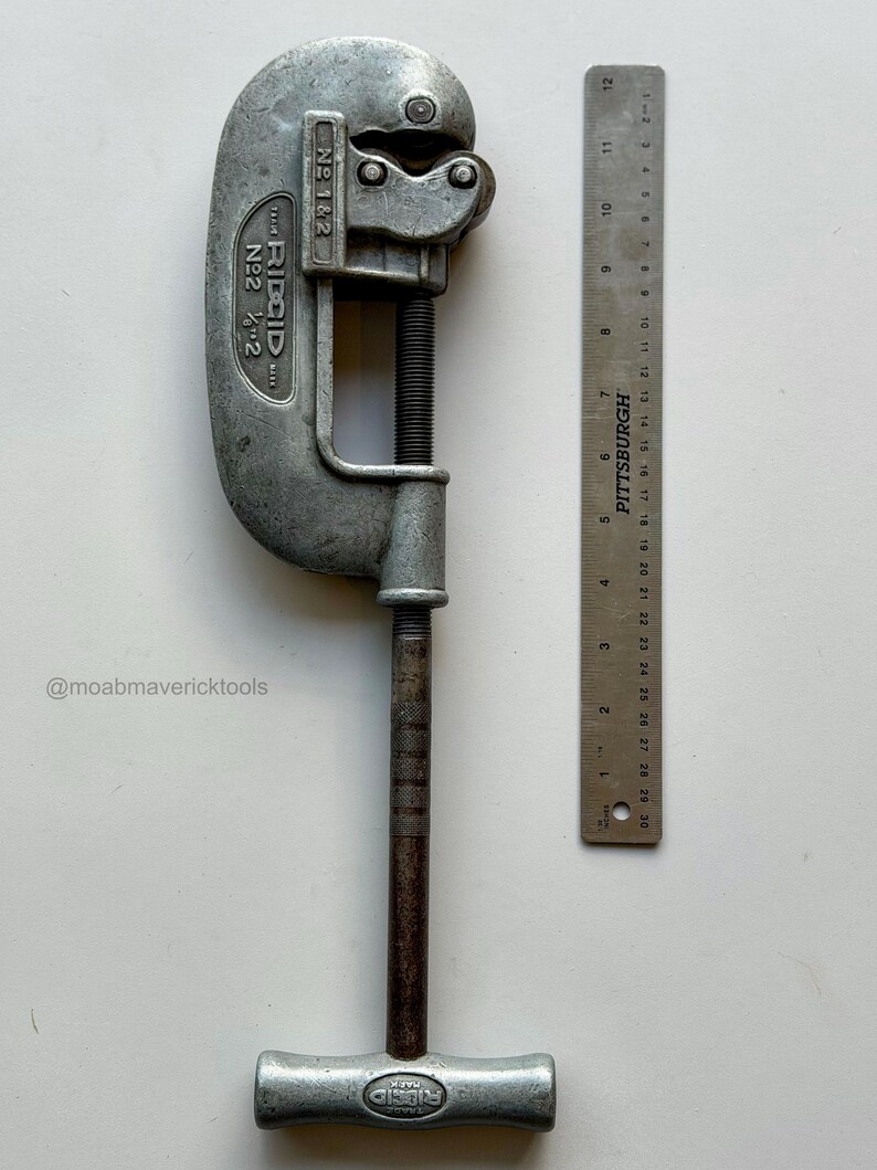 May include: A vintage RIDGID No. 2 pipe cutter, with a silver-colored body and a dark, textured handle. A Pittsburgh ruler, marked in inches, is positioned next to the cutter. The cutter is designed for pipes up to 2 inches.