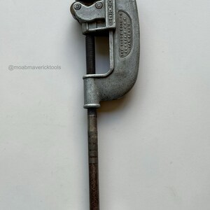May include: A vintage silver-colored pipe cutter tool. The tool has a curved body with a blade and a screw mechanism for adjusting the cutting depth. The handle is a T-shape. The text "RIDGID" is visible on the tool.