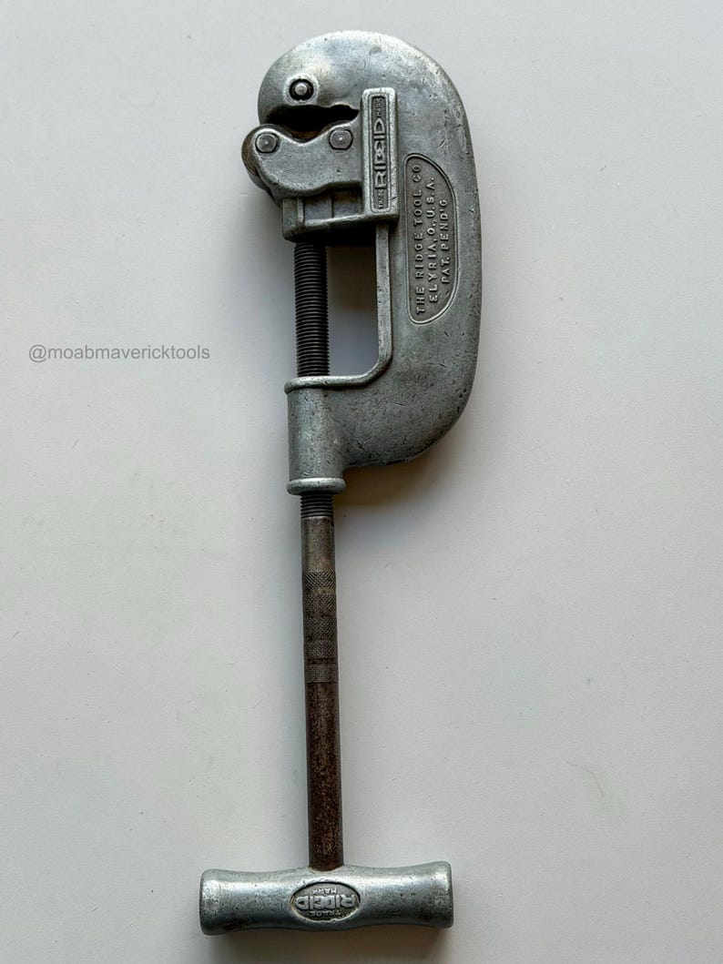 May include: A vintage silver-colored pipe cutter tool. The tool has a curved body with a blade and a screw mechanism for adjusting the cutting depth. The handle is a T-shape. The text "RIDGID" is visible on the tool.