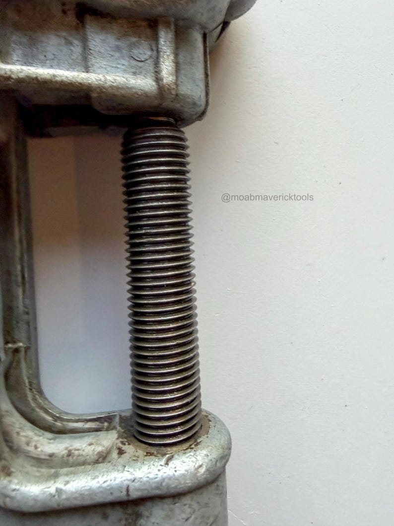 May include: Close-up of a vintage metal clamp with a threaded rod. The clamp is a dull silver color, showing signs of wear. The rod is tightly threaded, suggesting it's designed for securing objects. The image includes the text "@moabmavericktools".