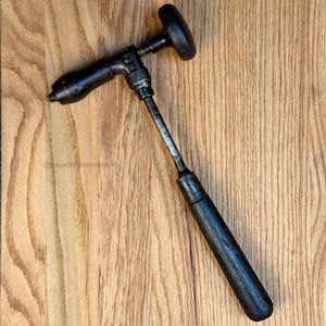 May include: A vintage hand drill with a black wooden handle and a metal body. The drill has a T-shaped top and a drill bit at the end. The tool is set against a wooden background.