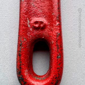 May include: Close-up of a red metal tool with a large oval opening. The tool has a textured, worn appearance with black markings, suggesting age and use. The red paint shows signs of wear.