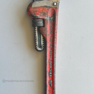 May include: A vintage red and silver pipe wrench, marked "THE RIDGE TOOL CO. ELYRIA, OHIO U.S.A. 10". The wrench shows signs of wear and tear, with a textured handle and adjustable jaws. The tool is approximately 10 inches in length.