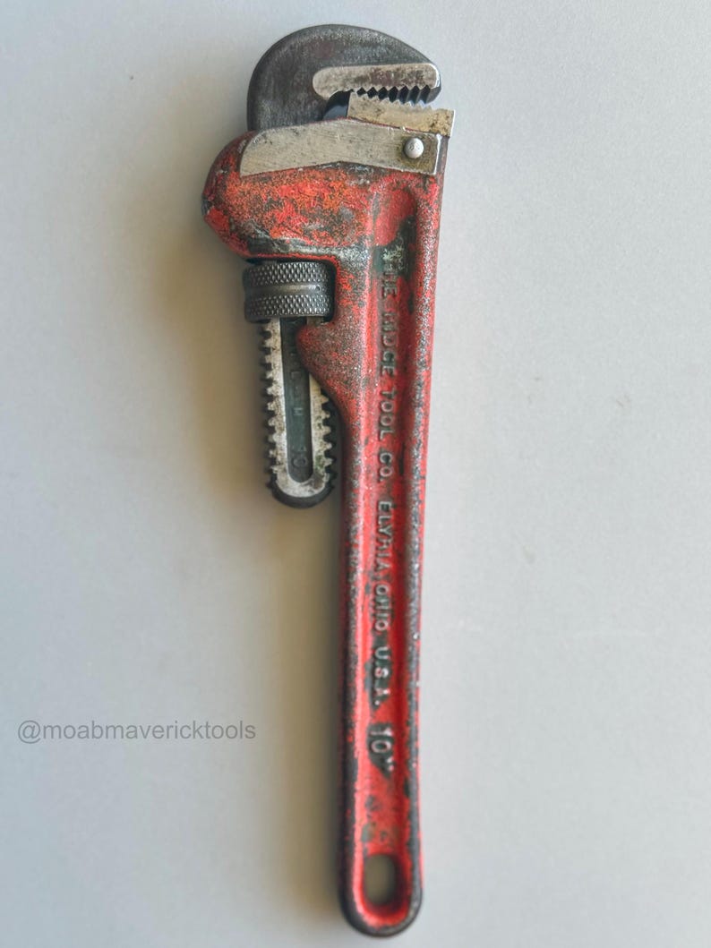 May include: A vintage red and silver pipe wrench, marked "THE RIDGE TOOL CO. ELYRIA, OHIO U.S.A. 10". The wrench shows signs of wear and tear, with a textured handle and adjustable jaws. The tool is approximately 10 inches in length.