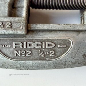 May include: Close-up of a vintage RIDGID No. 2 pipe cutter. The tool is made of silver metal and features the brand name and size specifications: 1/8 to 2 inches. The tool shows signs of wear, indicating its use.