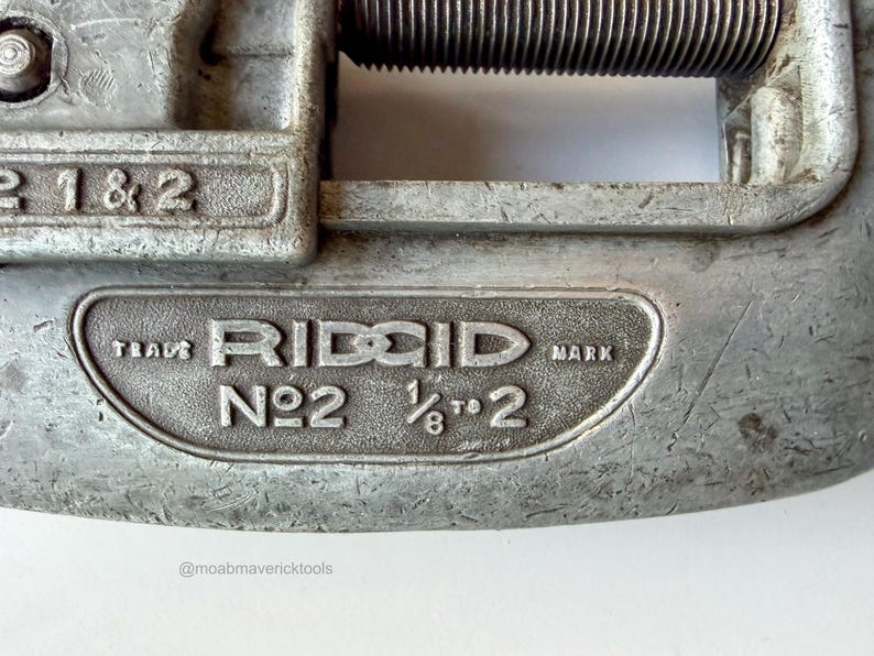 May include: Close-up of a vintage RIDGID No. 2 pipe cutter. The tool is made of silver metal and features the brand name and size specifications: 1/8 to 2 inches. The tool shows signs of wear, indicating its use.