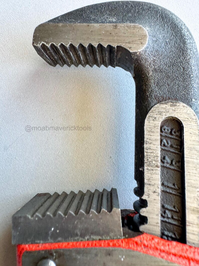 May include: Close-up of a heavy-duty pipe wrench. The wrench is made of dark gray metal with a red handle. The jaws have sharp, angled teeth for gripping pipes. The text "@moabmavericktools" is visible.