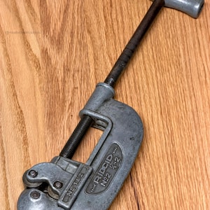 May include: A vintage Ridgid No. 2 1/2 pipe cutter tool, made of gray metal. The tool has a T-shaped handle and a screw mechanism for adjusting the cutting blade. The tool is designed to cut pipes up to 2 inches in diameter.