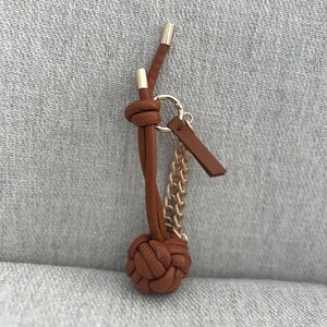 K&ouml;nnte beinhalten: Ein brauner Leder-Schl&uuml;sselanh&auml;nger mit geknotetem Design, goldfarbenen Akzenten und einer kleinen Kette. Der Schl&uuml;sselanh&auml;nger hat einen runden, gewebten Knoten an der Basis und ein kleines Lederetikett.