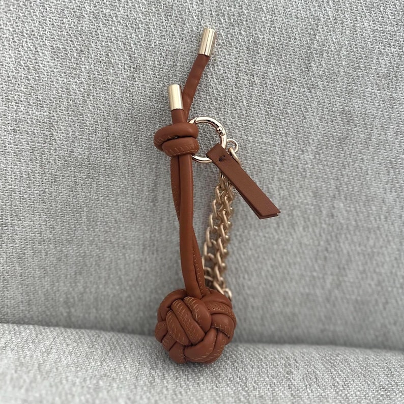 K&ouml;nnte beinhalten: Ein brauner Leder-Schl&uuml;sselanh&auml;nger mit geknotetem Design, goldfarbenen Akzenten und einer kleinen Kette. Der Schl&uuml;sselanh&auml;nger hat einen runden, gewebten Knoten an der Basis und ein kleines Lederetikett.