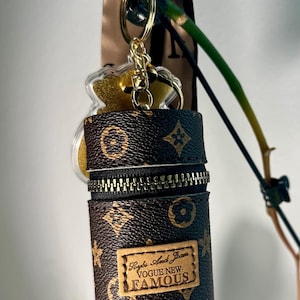 May include: A brown and tan cylindrical keychain case with a black zipper and gold hardware. The case has a repeating floral pattern and a rectangular label that reads "Paybs And Joan VOGUE NEW FAMOUS."