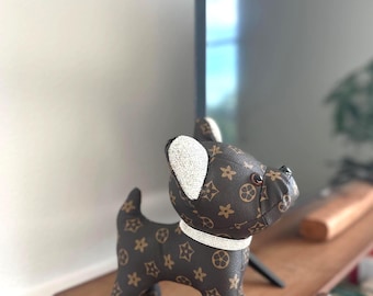 Könnte beinhalten: Braune und beige Hundefigur mit einem sich wiederholenden Muster aus Sternen und Kreisen. Der Hund hat ein weißes Halsband und weiße Akzente an den Ohren. Die Figur besteht aus einem lederartigen Material und steht auf einem Holzregal.