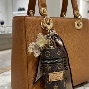 May include: A tan leather handbag with gold hardware, featuring a dangling charm set consisting of a clear acrylic teddy bear, a patterned silk ribbon, and a small cylindrical brown monogram pouch with the text Vogue New Famous.