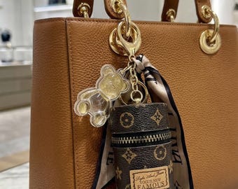 Könnte beinhalten: Eine hellbraune Ledertasche mit goldfarbenen Beschlägen, an der ein Anhänger-Set aus einem transparenten Acryl-Teddybären, einem gemusterten Seidenband und einer kleinen zylindrischen braunen Monogramm-Tasche mit dem Text Vogue New Famous hängt.