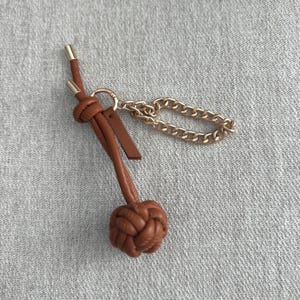 K&ouml;nnte beinhalten: Ein brauner Leder-Schl&uuml;sselanh&auml;nger mit dekorativem Knoten und goldfarbener Kette. Der Schl&uuml;sselanh&auml;nger hat unten ein geflochtenes Knotendesign und ein kleines Lederetikett. Die Kette ist kurz und klobig.
