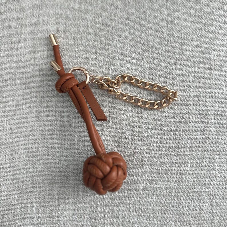 K&ouml;nnte beinhalten: Ein brauner Leder-Schl&uuml;sselanh&auml;nger mit dekorativem Knoten und goldfarbener Kette. Der Schl&uuml;sselanh&auml;nger hat unten ein geflochtenes Knotendesign und ein kleines Lederetikett. Die Kette ist kurz und klobig.