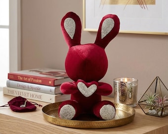 Könnte beinhalten: Ein rotes Samt-Hasen-Figürchen mit silbernen Akzenten an Ohren, Füßen und einem Herz. Der Hase steht auf einem goldenen Tablett, zusammen mit Büchern und einer Kerze, was eine dekorative Präsentation ergibt.